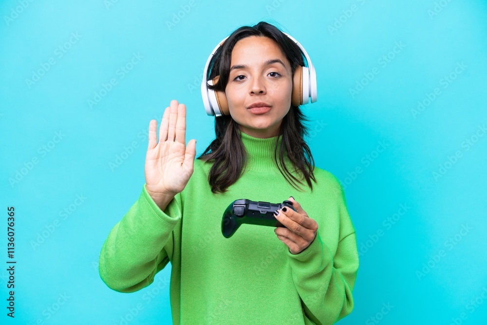 Young hispanic woman playing with a video game controller isolated on blue background making stop gesture Young hispanic woman playing with a video game controller isolated on blue background making stop gesture