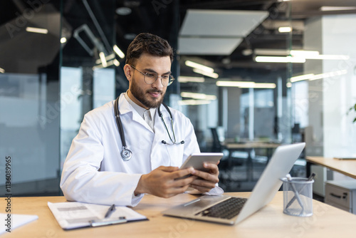 Mature man doctor seated at a desk, interacting with a tablet and laptop to access medical records, reflecting professional healthcare use of technology in a modern workspace.