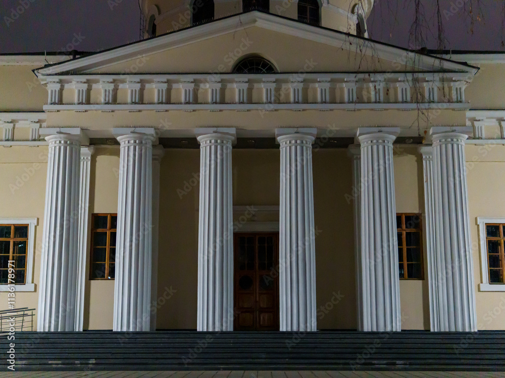 Fototapeta premium The strong and tall classical columns stand proud in the nighttime. The warm light accentuates the entrance of the building, creating a calm atmosphere in the surroundings.