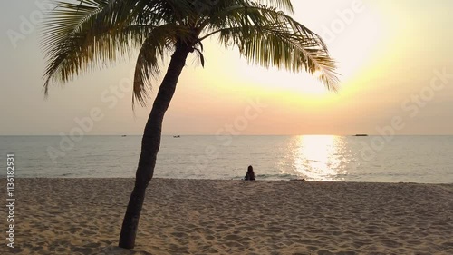 A lonely girl sits on the sandy beach under a palm tree and looks at the sun setting over the horizon
