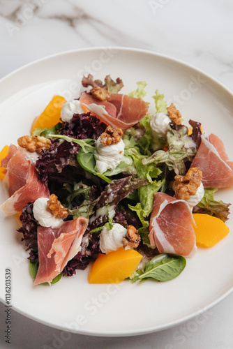Fresh salad with parma ham, melon slices, walnuts, goat cheese and mixed greens, served on a white plate on a marble table. Restaurant or cafe menu, cookbook recipe