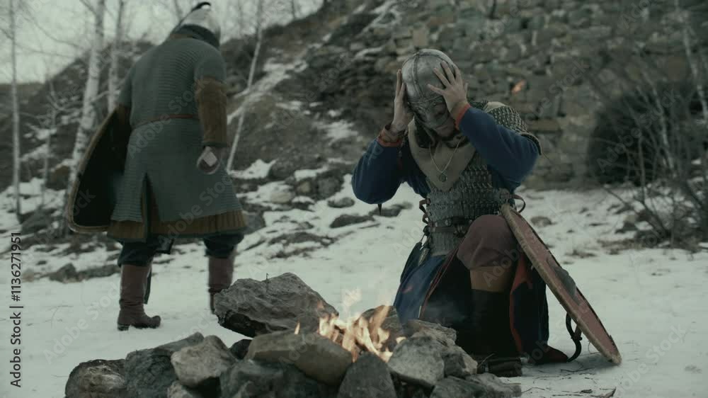 Medieval knight kneeling on snow-covered ground by fireplace, taking off helmet and warming his hands above flames in front of stone wall of fortress on cold winter day