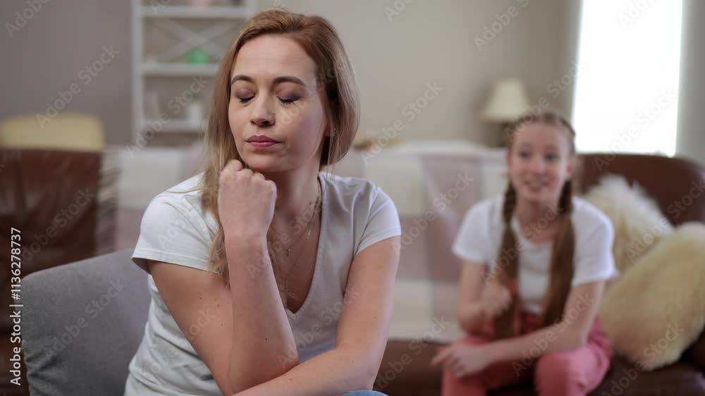A teenage girl animatedly gestures while sitting on the couch, attempting to explain something to her mother. The mother, sitting in the foreground with her chin resting on her hand, closes her eyes A teenage girl animatedly gestures while sitting on the couch, attempting to explain something to her mother. The mother, sitting in the foreground with her chin resting on her hand, closes her eyes
