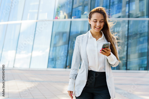 Woman in professional attire smiles while using a smartphone outside a modern glass building photo