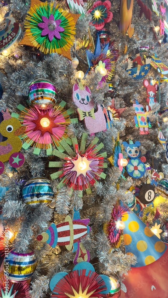 Obraz premium A silver Christmas tree adorned with vibrant ornaments, handcrafted flowers, festive hats, rockets, and toys like pink cats, robots, and yellow ducks. Perfect for holiday cards or banners.