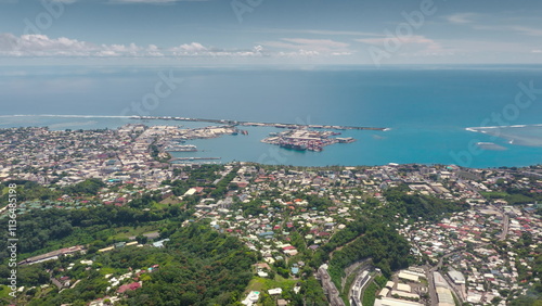 Aerial drone fly over Papeete coast, Tahiti cityscape harbor, green mountain hills, colorful village houses, streets, buildings, lush tropical vegetation against pacific ocean backdrop. Remote travel
