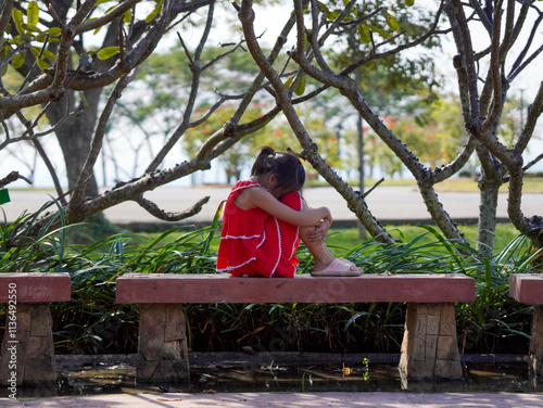 A sad little girl is sitting alone on bench in park. The little girl is unhappy and anxious because she has no one to play with. photo