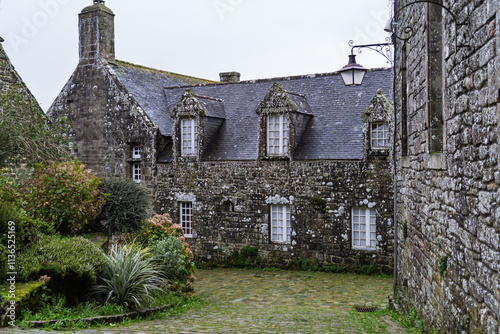 Une rue pavée de Locronan, les maisons anciennes aux fenêtres à petits carreaux et encadrement blanc se dressent sous un ciel gris. Les pavés mouillés ajoutent une touche de lumière douce à l'ensemble photo