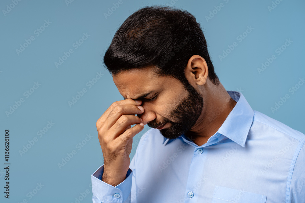 Worried Indian businessman holding head, eyes closed, showing pain and exhaustion on blue background Worried Indian businessman holding head, eyes closed, showing pain and exhaustion on blue background