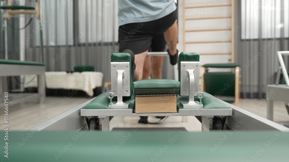 Man practicing kneeling stretch on pilates reformer with assistance to improve flexibility and strength. Trainer assist student doing lunge stretch on the reformer for enhancing balance. Habituate. Man practicing kneeling stretch on pilates reformer with assistance to improve flexibility and strength. Trainer assist student doing lunge stretch on the reformer for enhancing balance. Habituate.