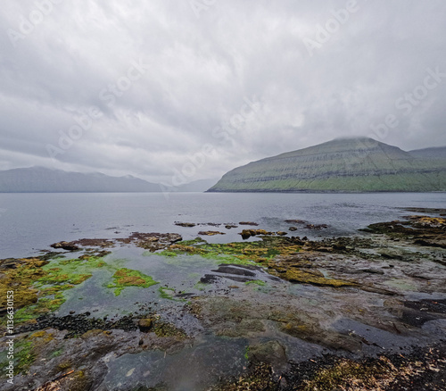 Dramatic low clouds hang over the majestic fjords on the island of Eysturoy, Faroe Islands. Rocky coastline meets the sea surrounded by lush green mountains. photo