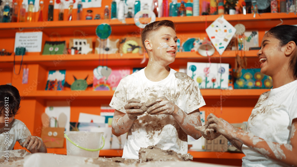 Group of happy diverse children playing clay while wearing muddy shirt at art lesson. Cute highschool girl playing with dough while putting clay on happy boy shirt at pottery workshop. Edification.