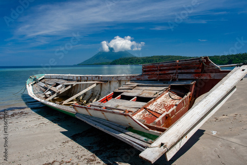 Old wooden boat stays on beach on Bunaken Island , Indonesia to get repairing and painting . On background we can see a vulcane with wait clouds , that  stopped over mountain and looks like a hat. photo