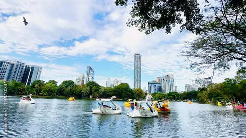 A vibrant city park scene with a tranquil lake surrounded by lush green trees and modern skyscrapers as a backdrop. In the foreground, two people in life jackets are playing in a duck boat a swan boat