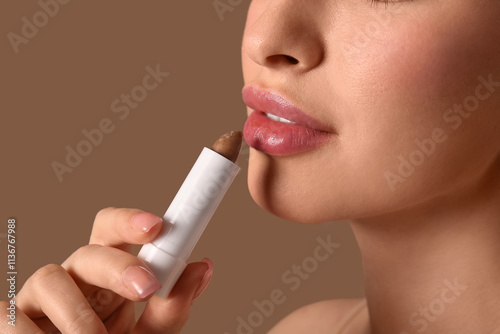 Young woman with lip balm on brown background, closeup