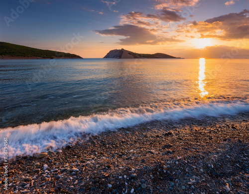 The sun sets dramatically over a mountain silhouette, casting golden reflections on the waves crashing onto a sandy beach. photo