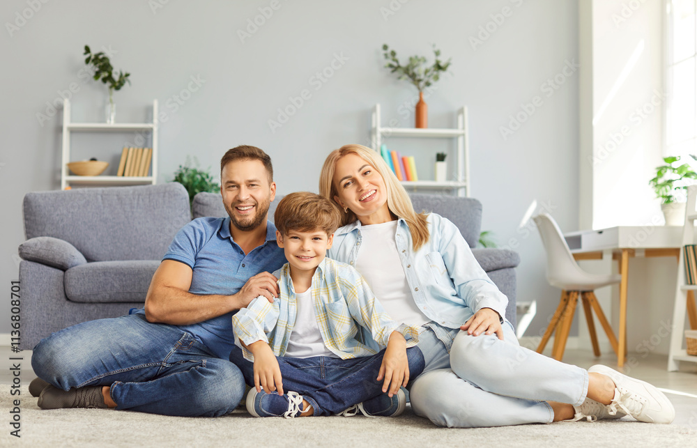 Happy parents and child making family portrait while sitting on floor in living room together. Smiling mother and father hugging their joyful son placing between them, looking at camera. Family bonds.