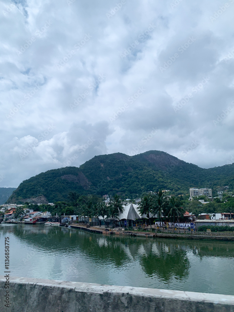 Scenic view of Rio de Janeiro with lush green mountains, calm river, palm trees, and cloudy sky. Scenic view of Rio de Janeiro with lush green mountains, calm river, palm trees, and cloudy sky.