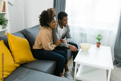 African American couple relaxes on a cozy sofa, sharing a joyful moment while watching a show with a bowl of popcorn. Essence of quality time and comfort in a stylish modern living room. Copy space