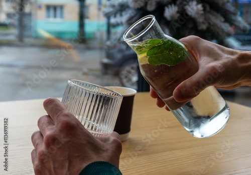 A person pours refreshing water with mint leaves from a carafe into a glass while seated at a cafe table during the morning