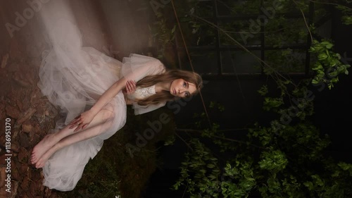 Caucasian woman with long hair in elegant white lace dress sits barefoot among dark foliage. Natural cosmetics or perfume concept.