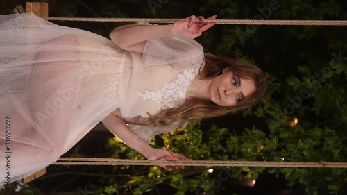 Caucasian woman with long hair in elegant white lace dress sits on wooden swing among dark foliage. Natural cosmetics or perfume concept.