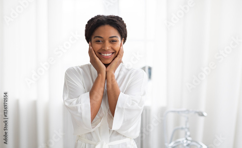 In a well-lit bathroom, a happy woman smiles as she participates in her skincare routine. Dressed in a soft robe, she radiates confidence and beauty while pampering herself.