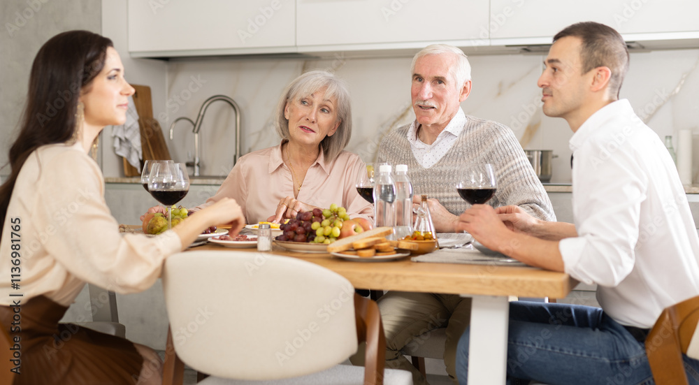 Two cheerful senior adults, man and woman, engaging in lively conversation with young couple at dinner table with wine and fruits in cozy home kitchen Two cheerful senior adults, man and woman, engaging in lively conversation with young couple at dinner table with wine and fruits in cozy home kitchen