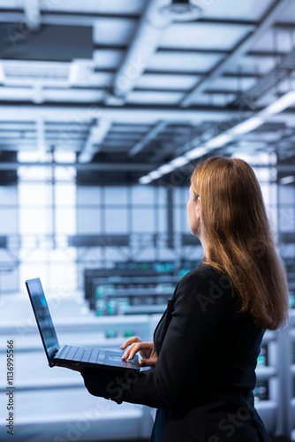 Software developer walking between server rows in data center, doing yearly infrastructure inspection. Worker in server farm establishment monitoring supercomputers equipment with laptop photo