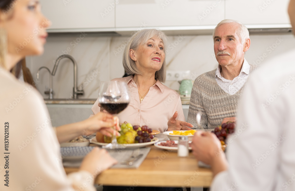 Mature spouses sitting at table, enjoying food and wine, chatting talking laughing with adult son and his girlfriend. In homely atmosphere male and female pensioner celebrate anniversary with guests