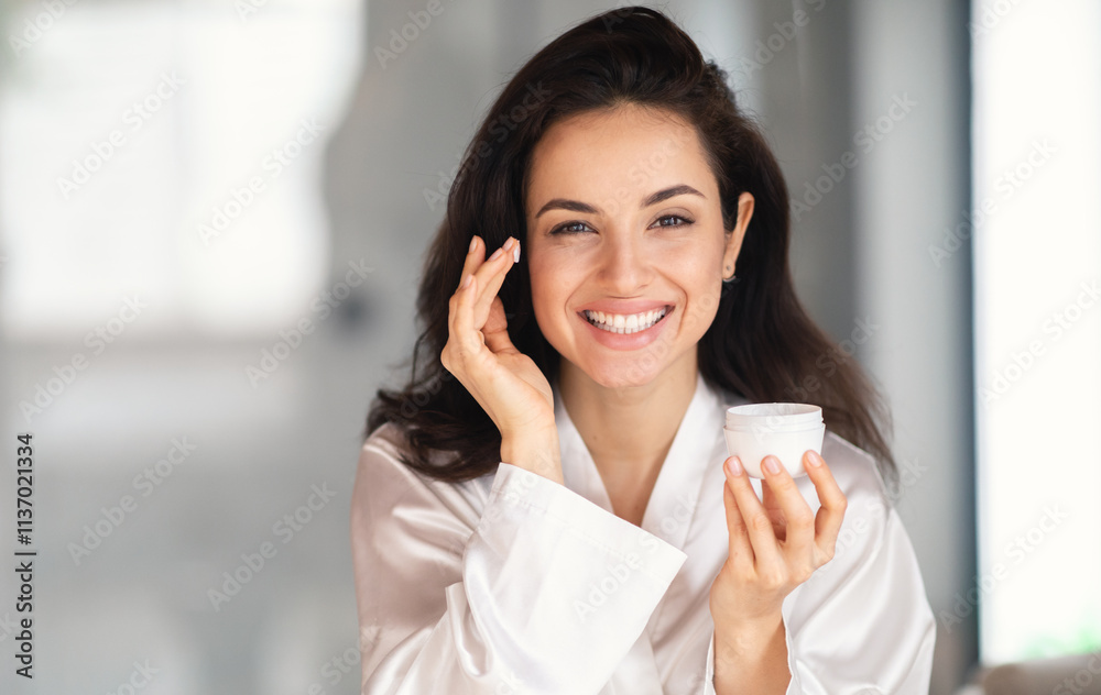 In a serene home bathroom, a young Caucasian brunette smiles while applying skin care cream. She radiates beauty and confidence in her pampering moment, surrounded by a relaxing atmosphere. In a serene home bathroom, a young Caucasian brunette smiles while applying skin care cream. She radiates beauty and confidence in her pampering moment, surrounded by a relaxing atmosphere.