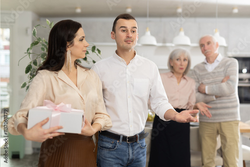 Positive interested young adult man gesturing welcomingly towards female guest holding gift box, against backdrop of disapproving elderly parents in domestic setting.... photo