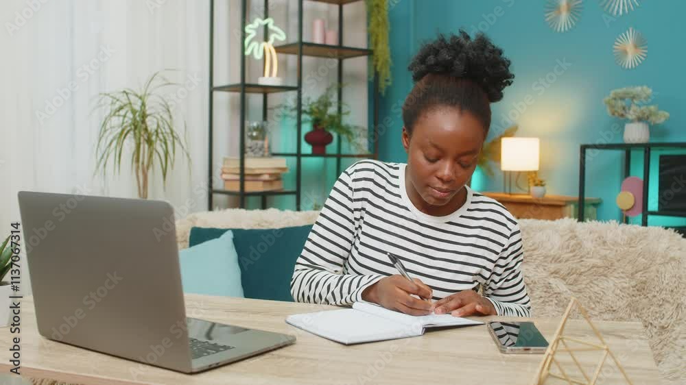 Afro-American Black woman working indoors at home. Young girl sitting on sofa using laptop, writing notes in notebook. Living room setting, focused expression, relaxed atmosphere, productivity. Afro-American Black woman working indoors at home. Young girl sitting on sofa using laptop, writing notes in notebook. Living room setting, focused expression, relaxed atmosphere, productivity.