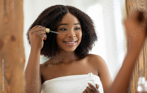 Beauty cosmetics for young skin. Sensual black woman applying regenerating serum onto her face near mirror indoors. Millennial African American lady pampering herself at home photo