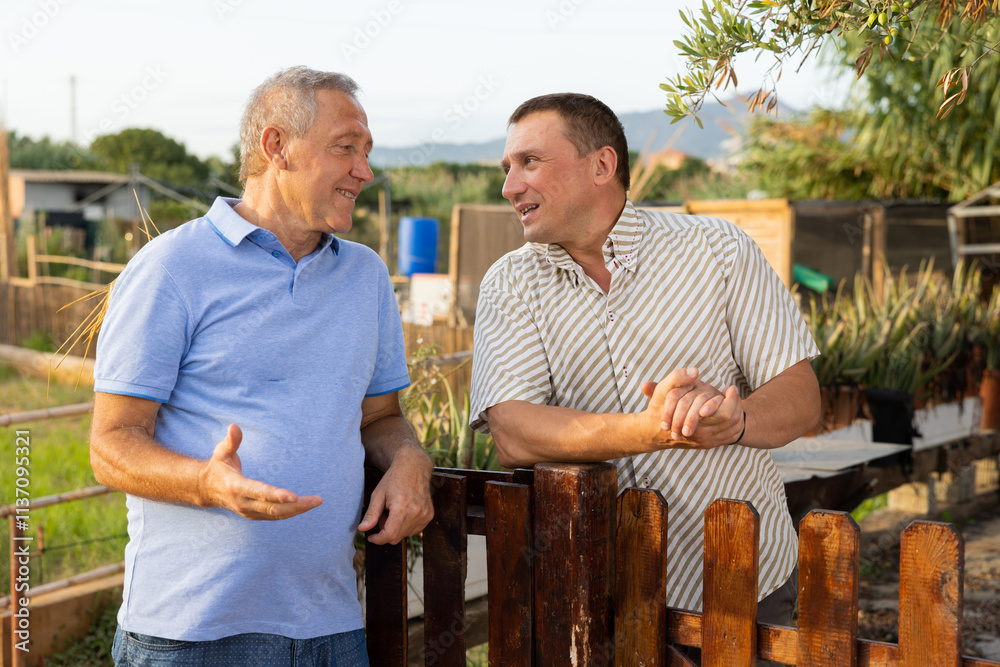 Two male gardeners talking near wooden fence in garden outdoor Two male gardeners talking near wooden fence in garden outdoor