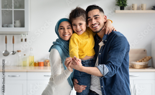 Portrait Of Happy Muslim Family Of Three With little Daughter Posing In Kitchen, Cheerful Islamic Father, Child And Mother In Hijab Embracing And Smiling At Camera, Bonding At Home Together