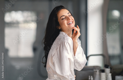 A young brunette woman smiles joyfully while pampering herself during a skincare routine at home. She relaxes in a contemporary bathroom, enhancing her beauty and self-care.