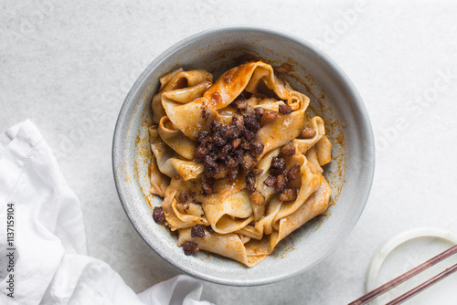 Overhead view of spicy wide noodles in a grey bowl, top view of biangbiang mian in a bowl, spicy belt noodles with green onions and diced pork in a bowl photo
