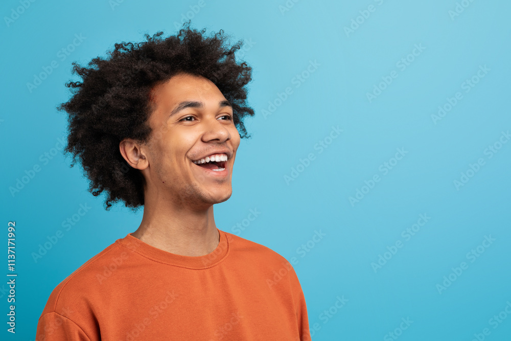 A young black man with curly hair laughs joyfully, exuding a sense of happiness. He is dressed casually in an orange sweater, set against a vibrant blue backdrop, copy space A young black man with curly hair laughs joyfully, exuding a sense of happiness. He is dressed casually in an orange sweater, set against a vibrant blue backdrop, copy space