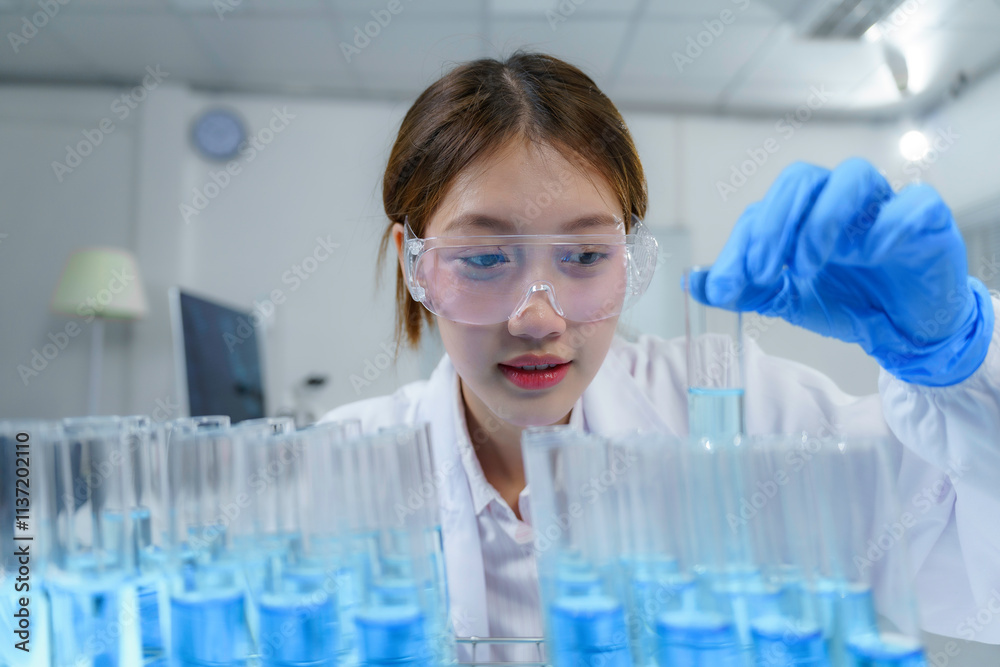 Young scientist wearing lab coat and protective glasses holding test tube analyzing blue liquid solution in modern laboratory Young scientist wearing lab coat and protective glasses holding test tube analyzing blue liquid solution in modern laboratory