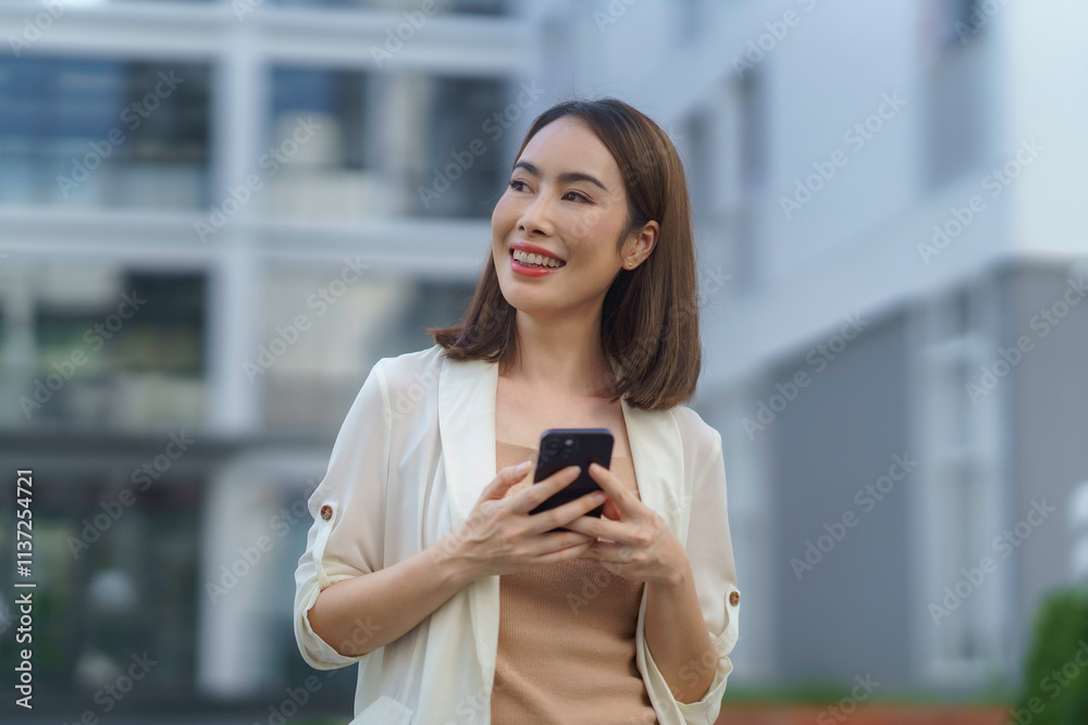Happy businesswoman is holding a smartphone and smiling while standing in front of a modern office building Happy businesswoman is holding a smartphone and smiling while standing in front of a modern office building
