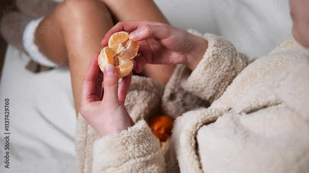 Woman in robe relaxes on white bed peeling mandarin orange while enjoying cozy atmosphere. She is wearing warm light socks and savoring every moment with mandarin orange concept of festive mood Woman in robe relaxes on white bed peeling mandarin orange while enjoying cozy atmosphere. She is wearing warm light socks and savoring every moment with mandarin orange concept of festive mood