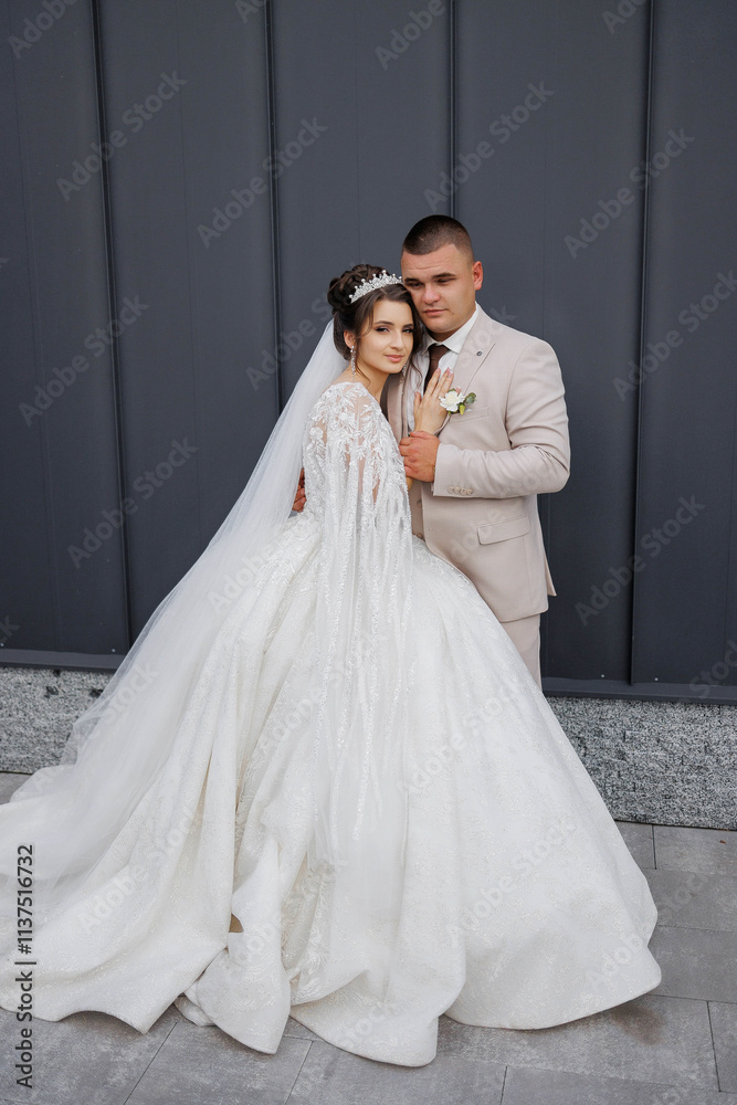 Fototapeta premium A bride and groom are posing for a picture in front of a gray wall. The bride is wearing a long white gown with a veil and a tiara, while the groom is wearing a suit