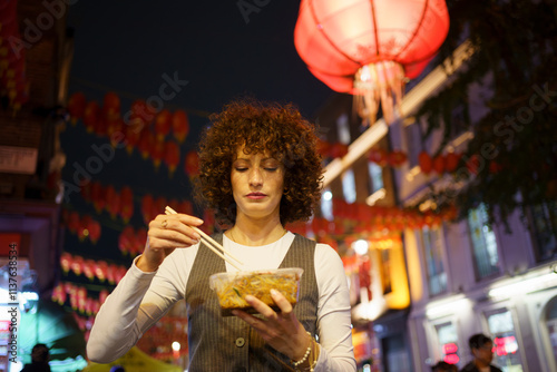 Curly haired woman eating noodles with chopsticks on street of Chinatown photo