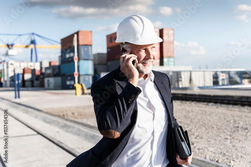 Businessman at cargo harbour, wearing safety helmet, using smartphone