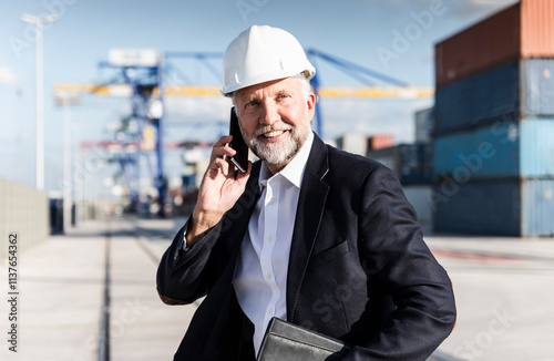 Businessman at cargo harbour, wearing safety helmet, using smartphone