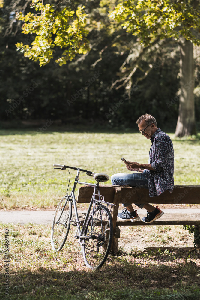 Senior man using mobile phone sitting on bench by bicycle in park Senior man using mobile phone sitting on bench by bicycle in park