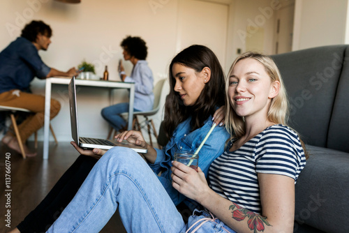 Two smiling young women sitting on floor sharing laptop with friends in background photo