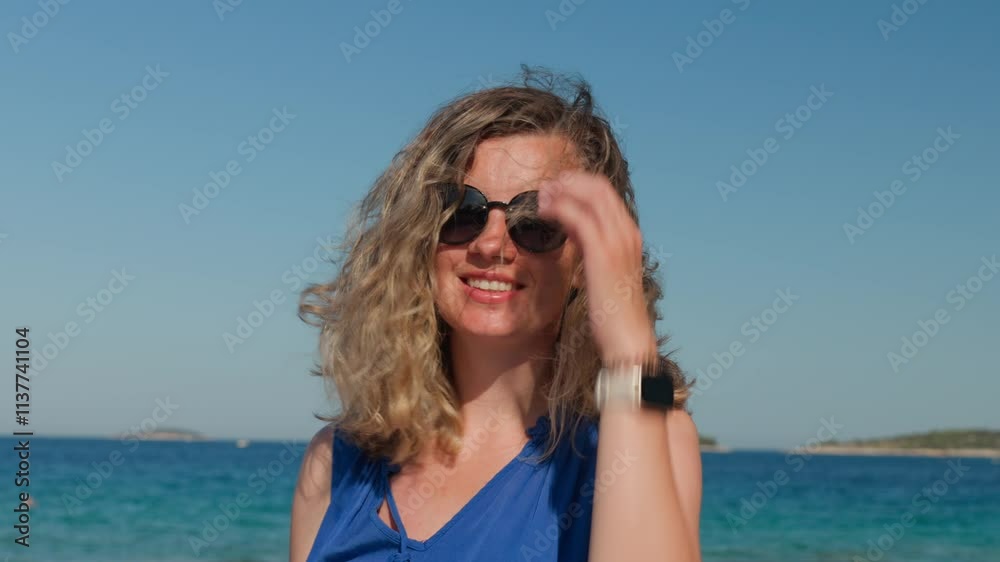 Woman in blue dress and sunglasses with flying hairs in windy day stands at sea coastline. Female tourist walking along sunny beach. Summer travel Woman in blue dress and sunglasses with flying hairs in windy day stands at sea coastline. Female tourist walking along sunny beach. Summer travel