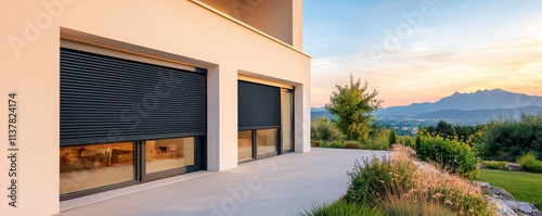 A modern home exterior featuring sleek roller shutters, a spacious patio, and a scenic view of mountains during sunset.